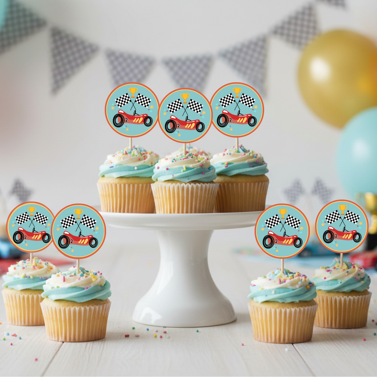 A set of racing-themed cupcake toppers displayed on cupcakes on a white pedestal stand, with a party background featuring balloons and bunting.